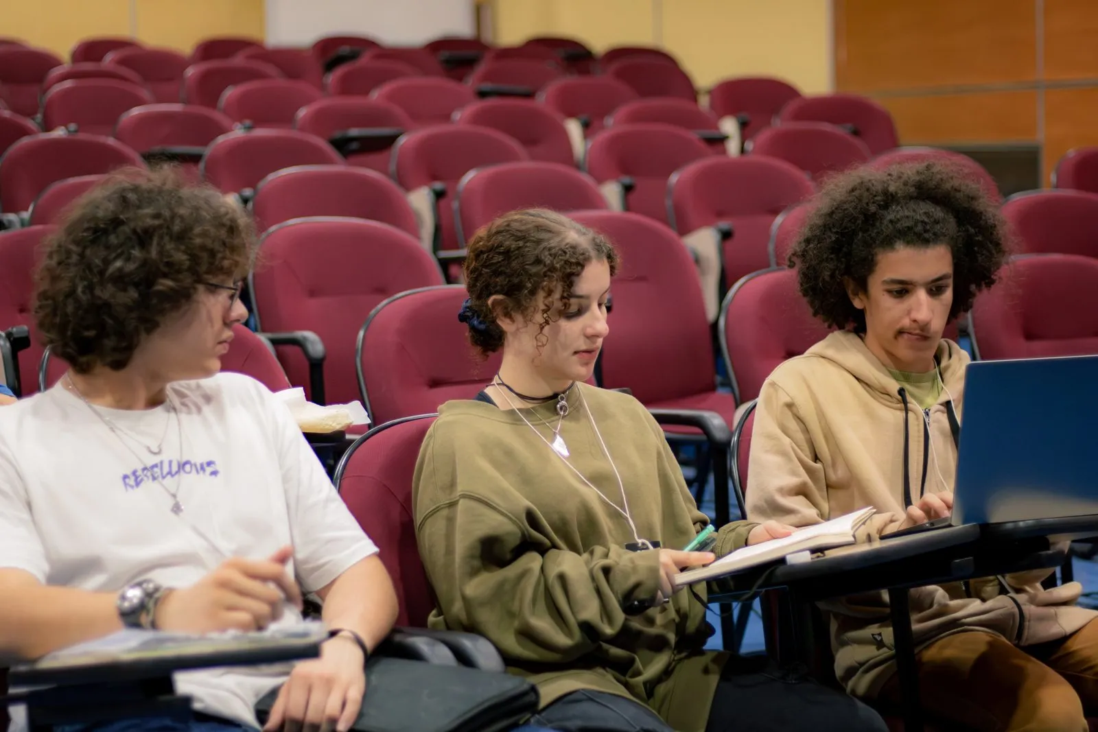 Three participants work with a notebook and a laptop in an auditorium.